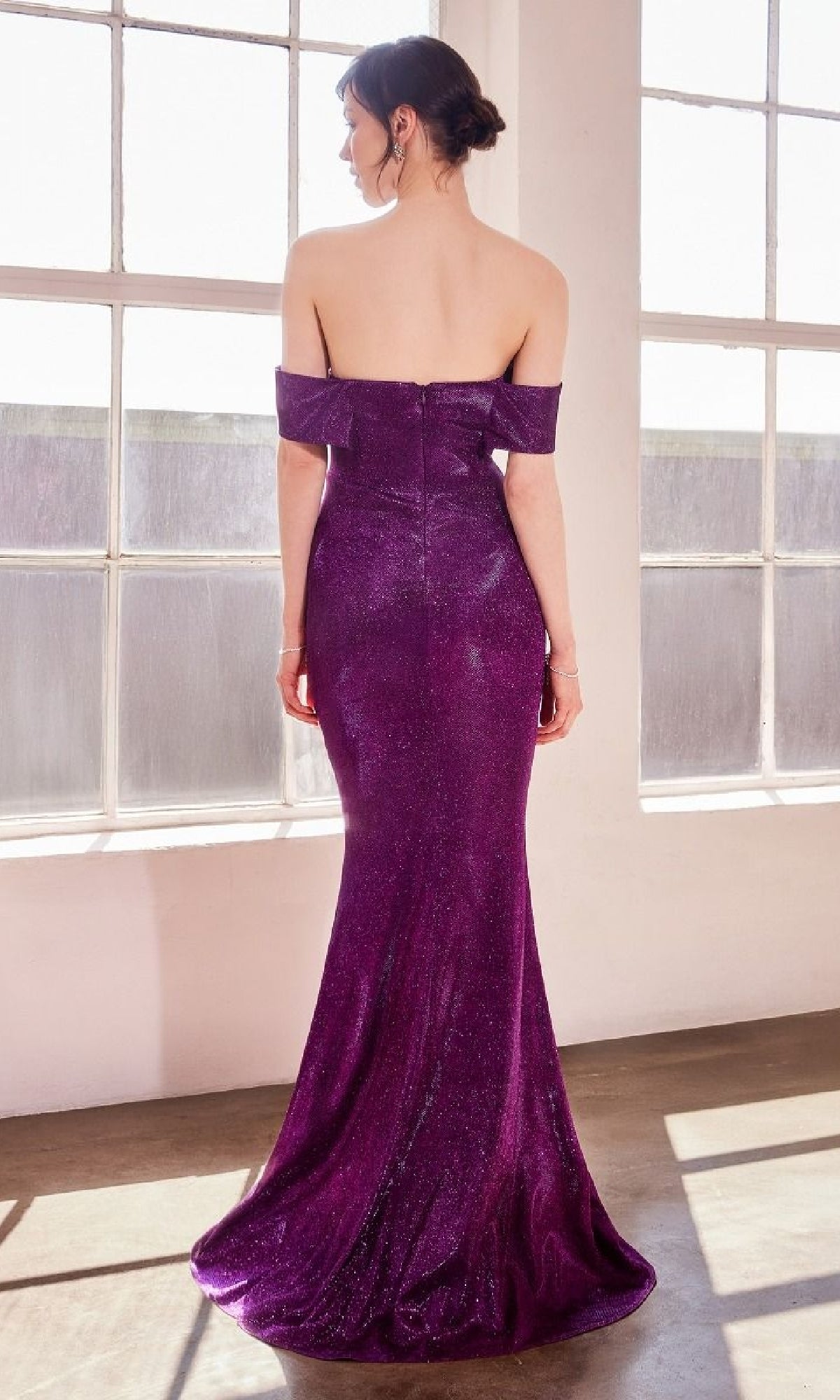 A woman in the Amoris T603 Off-Shoulder Long Glitter Prom Dress with a mermaid skirt and floor-length purple sequins stands indoors, her back to the camera as she faces large windows.